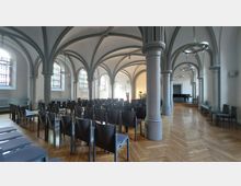 Innenansicht eines historischen Gebäudes mit gewölbter Decke aus grauen Bögen und tragenden Säulen. Der Raum hat hölzernen Fischgrätboden, Stuhlreihen, Buntglasfenster und im Hintergrund einen schwarzen Flügel sowie einen Altarbereich.