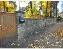 Steinmauer mit integriertem Holztor und einem schwarzen Briefkasten; im Hintergrund Herbstbäume mit gelbem Laub und ein Gebäude mit großen Fenstern. Der Boden ist mit gefallenen Blättern bedeckt, daneben ein unbefestigter Gehweg.