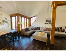 Modernes Wohnzimmer in einem Dachgeschoss mit Holzboden, großen Fenstern und einer Eckcouch mit bunten Kissen. Der Raum enthält einen Essbereich mit einem Holztisch, hängenden Pflanzen an der Wand und helle Holzbalken als gestalterische Elemente.