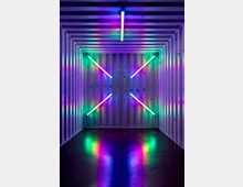 Ein rechteckiger Raum mit weißen, geriffelten Wänden und Decke, dekoriert mit bunten Leuchtstoffröhren in diagonaler Anordnung an der Rückwand und einer vertikalen Röhre an der Decke; die Lichtreflexionen erzeugen farbige Muster auf dem dunklen Boden.