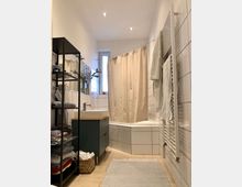 Badezimmer mit hellen Fliesenwänden, einer Eckbadewanne mit beigem Duschvorhang, einem kleinen Waschbecken auf einem grünen Unterschrank und einem offenen Regal mit Handtüchern. Ergänzt wird die Einrichtung durch einen Handtuchheizkörper und ein kleines Fenster im Hintergrund.