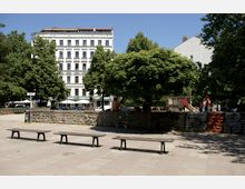 Städtischer Platz mit drei Holzsitzbänken im Vordergrund, umgeben von Bäumen und einem niedrigen Steinwall. Im Hintergrund befindet sich ein historisches weißes Wohngebäude mit Café-Terrasse, daneben ein kleiner Kinderspielbereich.