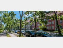 Straßenansicht mit Wohnhäusern in einer von Bäumen gesäumten Allee. Die Häuser haben rote und graue Fassaden mit weißen Fenstern, während am Straßenrand geparkte Autos und ein Gehweg sichtbar sind.