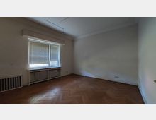 Ein leerer Raum mit weiß gestrichenen Wänden, einem großen Fenster mit heruntergelassenen Jalousien, darunter Heizkörper. Der Boden besteht aus Fischgrätparkett, und die Decke ist schlicht gestaltet.