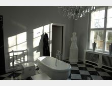 Helles Badezimmer mit freistehender Badewanne auf einem schwarz-weiß karierten Fliesenboden. Ein Kronleuchter hängt von der Decke, neben einem großen Fenster steht eine weiße Skulptur auf einem Sockel.