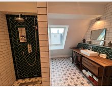 Modernes Badezimmer mit Dusche und Waschtisch. Die Dusche ist mit dunkelgrünen Fliesen im Fischgrätmuster verkleidet, während der Waschtisch aus Holz besteht und ein rechteckiges Waschbecken sowie ein Wandspiegel darüber hat. Der Boden ist mit gemusterten Fliesen gestaltet, und ein Dachfenster spendet Tageslicht.