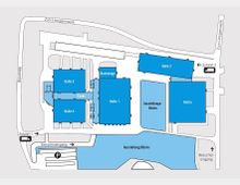 Übersichtskarte eines Veranstaltungsgeländes mit mehreren Gebäuden in blauer Farbe, darunter Halle 1 bis Halle 4, ein Foyer, ein Backstage-Bereich und ein Bistro. Neben den Gebäuden sind Ausstellungsflächen, Zufahrtswege, ein Besucherparkplatz und Eingänge eingezeichnet.