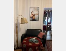 Kleiner Sitzbereich in einem gemütlichen Innenraum mit einer schwarzen Ledercouch, einem runden Tisch mit rotem Blumen-Tischtuch und einer Stehlampe im Retro-Stil. Ein gerahmtes Bild eines Bogenschützen hängt an der weißen Wand, im Hintergrund sind Holzbalken und weitere Möbelelemente erkennbar.