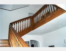 Innenansicht eines Treppenhauses mit einer Holztreppe und einem Geländer aus dunklem Holz. Im Hintergrund ist eine weiße Wand mit einem rundbogigen Durchgang zu sehen.