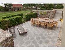 Steinterrasse mit Holzmöbeln, darunter ein langer Tisch mit mehreren Stühlen und zwei Liegen, umgeben von niedrigen Hecken und einer Natursteinmauer. Im Hintergrund befindet sich eine Wiese mit jungen Bäumen sowie Gebäuden mit roten Ziegeldächern.