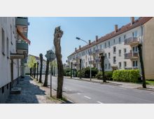 Straßenansicht mit Wohnhäusern im Hintergrund sowie einer mehrspurigen Straße und einem Gehweg. Die Fassaden der Häuser sind hell, mit Balkonen und roten Dächern, während zurückgeschnittene Bäume und Büsche den Straßenrand säumen.