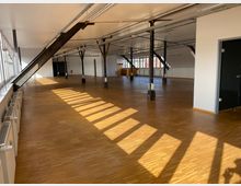 Großer, offener Innenraum mit heller Holzbodenfläche, schrägen Stahlträgern, sichtbaren Deckenverkabelungen und großen Fenstern, die das Tageslicht hereinlassen. Der Raum hat freistehende Säulen und weiße Wände, die ein modernes, industrielles Ambiente schaffen.