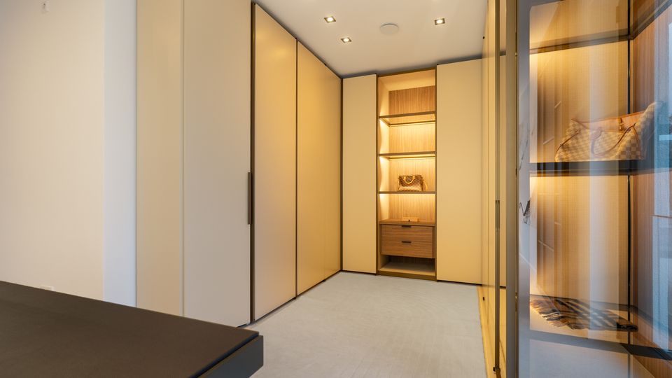 Moderner begehbarer Kleiderschrank mit deckenhohen, cremefarbenen Einbauschränken und einem beleuchteten Holzregal mit Schublade in der Mitte. Rechts sind gläserne Regaleinsätze mit integrierter Beleuchtung und schwarze Rahmen zu sehen, darüber eingelassene Decken-Spots.