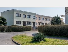 Reihenhäuser mit flachen Dächern und pastellfarbenen Fassaden, darunter Fenster und Eingänge mit kleinen Treppenaufgängen, umgeben von niedrigen Hecken und Grünflächen. Im Vordergrund ist ein Gehweg und gepflegter Rasen mit Pflanzen sichtbar.