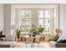 Wohnzimmer mit großen, weißen Sprossenfenstern, durch die Bäume und ein Wohngebäude im Hintergrund sichtbar sind. Zentral steht ein runder Tisch mit vier Stühlen, umgeben von Zimmerpflanzen, zwei grauen Sofas und dezenten Lampen; Holzboden und helle Wände schaffen eine einladende Atmosphäre.