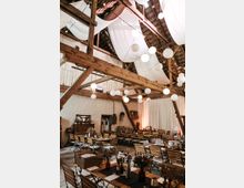 Ein rustikales Veranstaltungsgebäude mit sichtbaren Holzbalken und einer hohen Decke, dekoriert mit weißen Stoffbahnen und hängenden Papierlampen. Der Raum ist mit Holztischen und -stühlen eingerichtet, die für ein Fest oder eine Feier vorbereitet sind, mit Blumen und Tischdekorationen.
