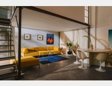 Modernes Loft-Wohnzimmer mit Zwischenebene und schwarzer Metalltreppe, Betonboden und hoher Fensterfront mit Vorhängen. Gelbes Ecksofa mit Glastisch auf blauem Teppich neben einem runden Esstisch mit weißen Tulpenstühlen, dazu Wandkunst und Zimmerpflanzen.