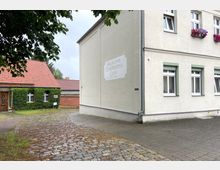 Straßenszene mit einem hellen Gebäude auf der rechten Seite, das „Bäckerei, Conditorei & Café“ zeigt, und einem kleinen Haus mit grün bewachsener Fassade und rotem Ziegeldach auf der linken Seite. Der Boden besteht aus Kopfsteinpflaster und gepflasterten Wegen, umgeben von einigen Bäumen.