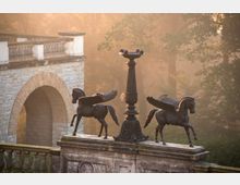 Steinbrücke mit verziertem Geländer, das zwei Skulpturen geflügelter Pferde und eine dekorative Laterne zeigt, umgeben von Bäumen in nebligem Morgenlicht. Im Hintergrund ist ein großes gemauertes Rundbogenportal zu sehen.