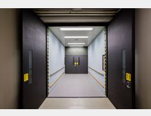 Großer Industrieaufzug mit geöffneten schwarzen Schiebetüren, gelb-schwarzen Warnmarkierungen an den Türrahmen, hellblauen Wänden, Holzhandläufen und einer Doppeltür am Ende des Aufzugs. Die Decke ist mit rechteckigen LED-Leuchten ausgestattet.