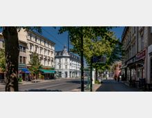 Straßenszene einer Stadt mit mehrstöckigen Gebäuden im historischen Stil, darunter Geschäfte und Restaurants mit Markisen und Schildern. Im Vordergrund befinden sich eine Baumreihe, ein Gehweg mit Passanten und eine Bushaltestelle mit Fahrplananzeige.