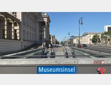Eingang zur U-Bahn-Station „Museumsinsel“ mit glasüberdachten Rolltreppen, flankiert von historischen Gebäuden mit Säulenfassaden entlang einer breiten Straße. Im Hintergrund sind Laternen im klassischen Stil und weitere Stadtarchitektur sichtbar.