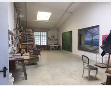 Atelier mit hoher Decke und Oberlicht, ausgestattet mit Arbeitsflächen, Regalen voller Materialien und Werkzeugen sowie großen Gemälden an der rechten Wand; im Hintergrund ein Fenster mit Milchglas und weitere Möbel.