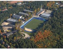 Luftaufnahme eines Sportkomplexes mit einem Fußballplatz, umgeben von einer blauen Laufbahn. Der Komplex wird von mehreren Gebäuden und dichten Waldgebieten umgeben, teils in herbstlichen Farben.