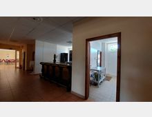 Innenansicht eines Gebäudes mit einem gefliesten Boden und einer Decke aus weißen Paneelen. Links führt ein Gang in einen angrenzenden Veranstaltungsraum mit Stuhlreihen, während rechts eine Tür in eine kleine Küche mit Edelstahl-Arbeitsflächen und weiß gefliesten Wänden führt.