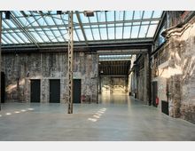 Großer, lichtdurchfluteter Raum in einem Industriegebäude mit Backsteinwänden, einem Boden aus poliertem Beton und einem Glasdach. Die Wände sind mit mehreren schwarzen Türen und sichtbaren Rohren versehen, während die Decke von Metallträgern gestützt wird.