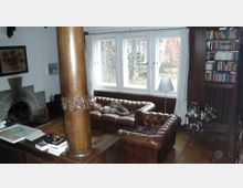 Wohnzimmer mit klassischen Elementen, darunter Ledersofas in Chesterfield-Stil, ein Kamin mit Rundbogen, eine verzierte Holzsäule und eine Fensterfront mit weißen Vorhängen. Im Hintergrund ein Bücherregal und eine Bar-Ecke mit Flaschen.