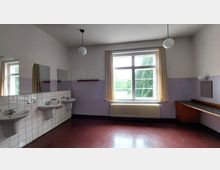 Ein einfacher Waschraum mit drei weißen Waschbecken und Spiegeln auf der linken Seite, weiß gefliesten Wänden und einem großen, halboffenen Fenster mit cremefarbenem Vorhang. Rechts befinden sich ein Heizkörper und eine Holzablage, der Boden ist rotbraun, und zwei Kugellampen hängen von der Decke.