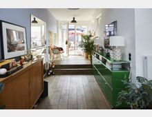 Wohnbereich mit modernem Interieur: Ein schmaler Raum mit Holzfußboden, einer Holzkommode auf der linken Seite und einem grünen Metallschrank auf der rechten. Große Glastüren im Hintergrund führen zu einer Terrasse mit Sonnenschirm, und mehrere Pflanzen sowie Dekorationsgegenstände ergänzen das Design.