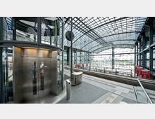 Innenansicht eines modernen Bahnhofs mit einem gläsernen Aufzug auf der linken Seite und einer weitläufigen Bahnsteighalle mit gebogener Glasdachkonstruktion. Im Hintergrund sind Gleise, Beschilderungen und helle Außenbereiche erkennbar.