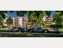 Spielplatz mit Klettergerüst und wellenförmiger Mauer in einer parkähnlichen Umgebung, umgeben von Bäumen. Im Hintergrund befinden sich mehrstöckige Wohngebäude mit bunten Balkonen.