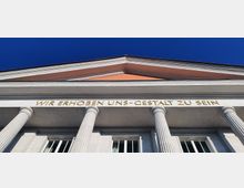 Fassade eines neoklassizistischen Gebäudes mit vier Säulen und einem Giebel; über den Fenstern steht in goldenen Buchstaben der Schriftzug "WIR ERHOBEN UNS - GESTALT ZU SEIN". Der Himmel ist klar und blau.