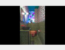 Kleines Badezimmer mit auffälliger Inneneinrichtung: die unteren Wände bestehen aus einem dunkelgrünen Terrazzo-Material, während die oberen Bereiche mit rosa gemustertem Tapetendesign dekoriert sind. Ein farbiges Glassteinfenster lässt diffuses Licht herein, und das Waschbecken sowie die Toilette sind in modernen Formen und kräftigen Farben gestaltet.