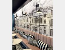 Langer Sitzbereich in einem Café oder Restaurant mit gepolsterten rosa Bänken, gestreiften und geometrisch gemusterten Kissen. An der Wand ist eine großflächige schwarz-weiße Illustration historischer Gebäude zu sehen, beleuchtet von modernen, schwarzen Wandlampen.