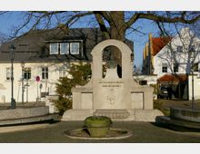 Denkmal mit einer Büste in einem steinernen Bogen und Inschrift, gewidmet dem Schöpfer des Teltowkanals, umgeben von einer kleinen Brunnenanlage und einem Baum; im Hintergrund stehen helle Wohnhäuser mit roten Dächern und Laternen.