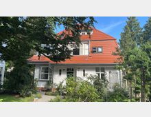 Zweistöckiges Wohnhaus mit weißer Fassade und einem roten Giebeldach, umgeben von einem Garten mit Bäumen und Sträuchern. Ein kleiner Weg führt zur Haustür in der Mitte des Gebäudes.