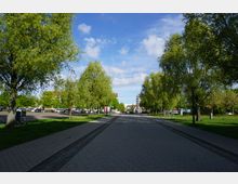 Eine gepflasterte Fußgängerzone mit Baumreihen auf beiden Seiten führt durch einen parkähnlichen Bereich. Im Hintergrund sind parkende Autos, Gebäude und ein teilweise eingerüstetes Bauwerk zu sehen.