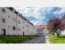 Wohnhof mit mehrstöckigen, hellen Reihenwohngebäuden und roten Dächern, flankiert von gepflegten Rasenflächen und einem blühenden Baum. Ein gepflasterter Weg führt zwischen den Häusern entlang, parkende Autos sind im Hintergrund sichtbar.