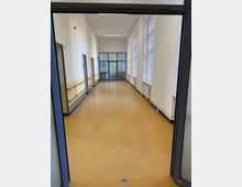 Ein heller Flur mit gelb-beigem Bodenbelag, weißen Wänden und mehreren Fenstern auf der rechten Seite. Auf der linken Seite befinden sich Türen und eine gelb abgesetzte Wand mit Handläufen; am Ende des Flurs ist eine Glastür zu sehen.