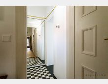 Heller Flur in einer Wohnung mit weißen Wänden, schwarz-weiß kariertem Boden und einer großen Wandspiegelfläche, die den Raum optisch vergrößert. Im Hintergrund führt der Flur zu einem Bereich mit Holzboden und einer Tür mit Glasfenstern.