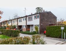 Mehrfamilienhäuser in einer Reihenhaussiedlung mit zweigeschossigen Gebäuden, abwechselnd verklinkert und mit hellen Fassadenelementen. Im Vordergrund befinden sich Hecken, gepflegte Vorgärten sowie ein Schild mit der Aufschrift „Privatstraße“.