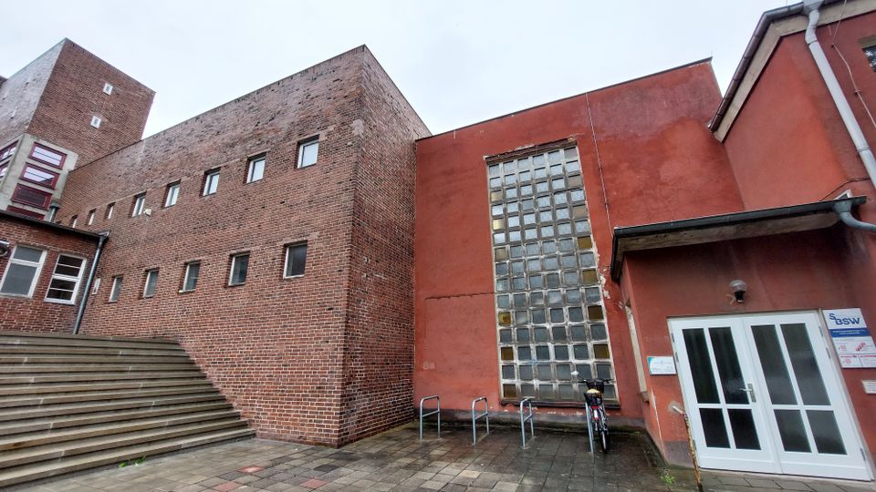 Backstein-Gebäudefassade mit kleinen rechteckigen Fenstern, neben einem roten Gebäudeteil mit einer großen Glasbausteinwand. Im Vordergrund führen steinerne Treppenstufen auf einen gepflasterten Platz, auf dem Fahrradständer und eine Tür mit Glasfenstern sichtbar sind.
