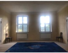 Ein schlichter Raum mit zwei großen Fenstern, hellgelben Wänden und hellem Holzboden. Im Vordergrund liegt ein großes blaues Teppichmuster, und an den Seiten stehen zwei Holzstühle an den Wänden.