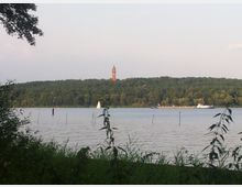 Blick auf einen See mit einer bewaldeten Hügellandschaft im Hintergrund; auf einem Hügel steht ein hoher, roter Turm. Im Vordergrund befinden sich Gräser und Sträucher, auf dem Wasser segelt ein kleines Boot, und ein Frachtschiff ist im Hintergrund zu sehen.