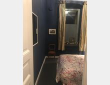 Kleines Schlafzimmer mit dunkelblauen Wänden, einem Bett mit gemusterter Tagesdecke und einem Fenster mit hellen Vorhängen über einem Heizkörper. An der Wand hängen ein Spiegel und ein Bild, daneben steht ein kleiner Holzstuhl; auf der Fensterbank liegen Dekoobjekte.