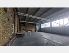 Industriehalle mit Backsteinwänden, großflächigen Fensterfronten mit Metallverstrebungen und einer Decke aus Sichtbeton. Im Raum stehen einfache Tische und Stühle auf einem Betonboden.